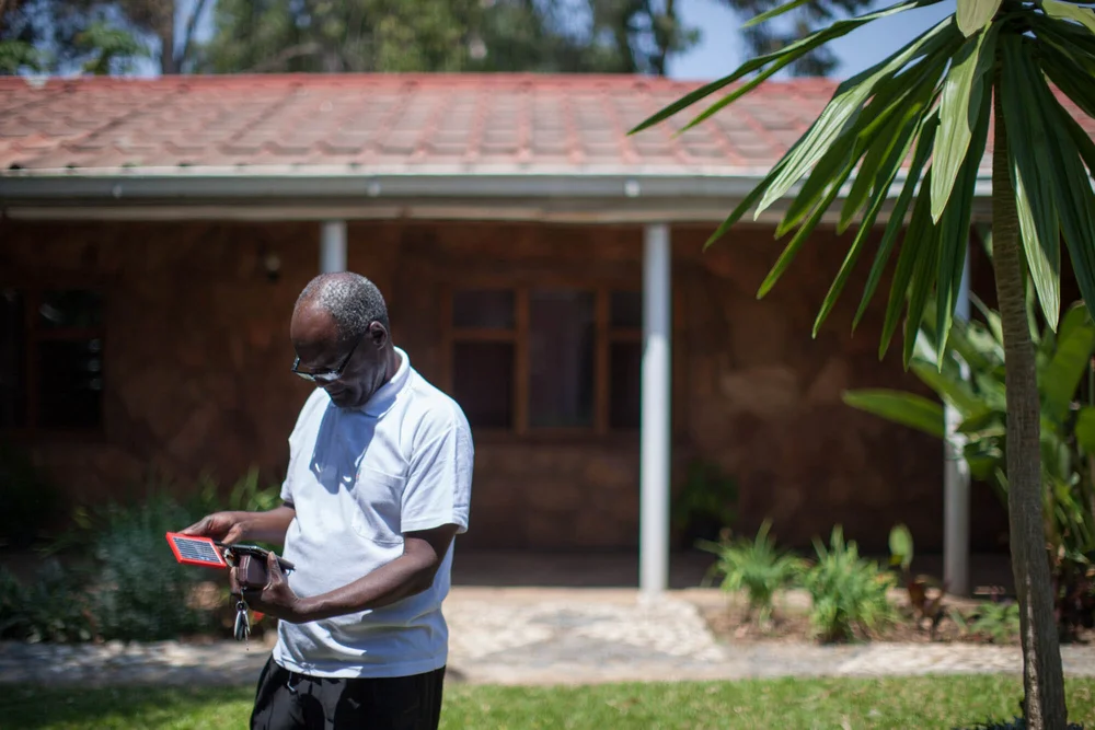 An older man in a white polo shirt examining a small red-bordered solar device on a lawn in front of a stone house with a covered porch