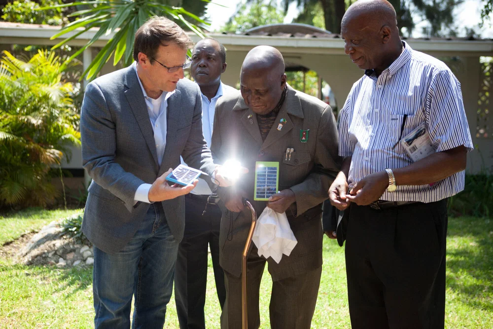 Four men on a sunlit lawn examining compact World Panel solar charging panels, one tilting sunlight toward the camera