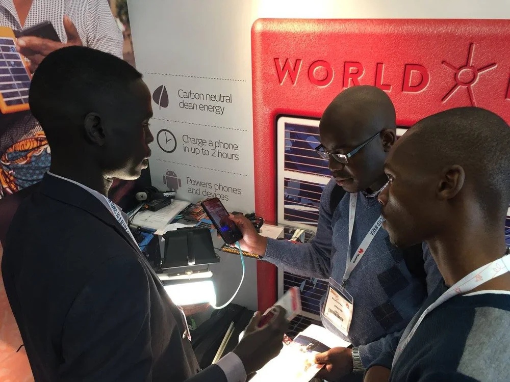 Three men at the World Panel trade show booth examining a phone on a teal charging cable, beside wall points reading “Carbon neutral clean energy”, “Charge a phone in up to 2 hours” and “Powers phones and devices”