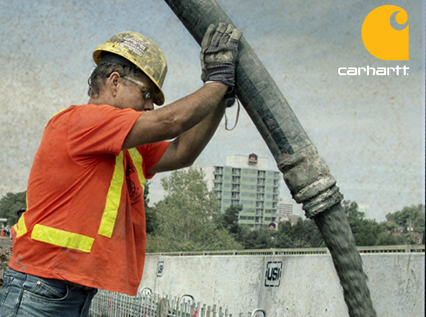 Carhartt campaign ad: a worker in a yellow hardhat and orange high-visibility shirt guiding a concrete pump hose