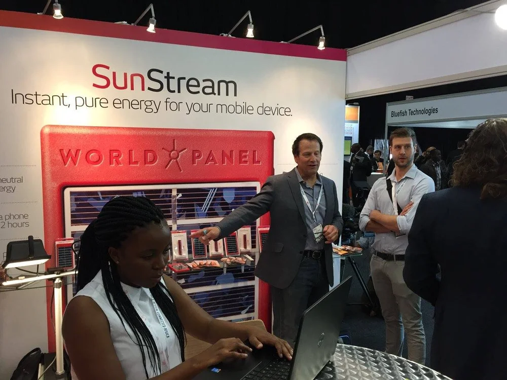 World Panel trade show booth: a woman at a laptop in front of a SunStream display carrying the tagline “Instant, pure energy for your mobile device.”, with two men behind the counter