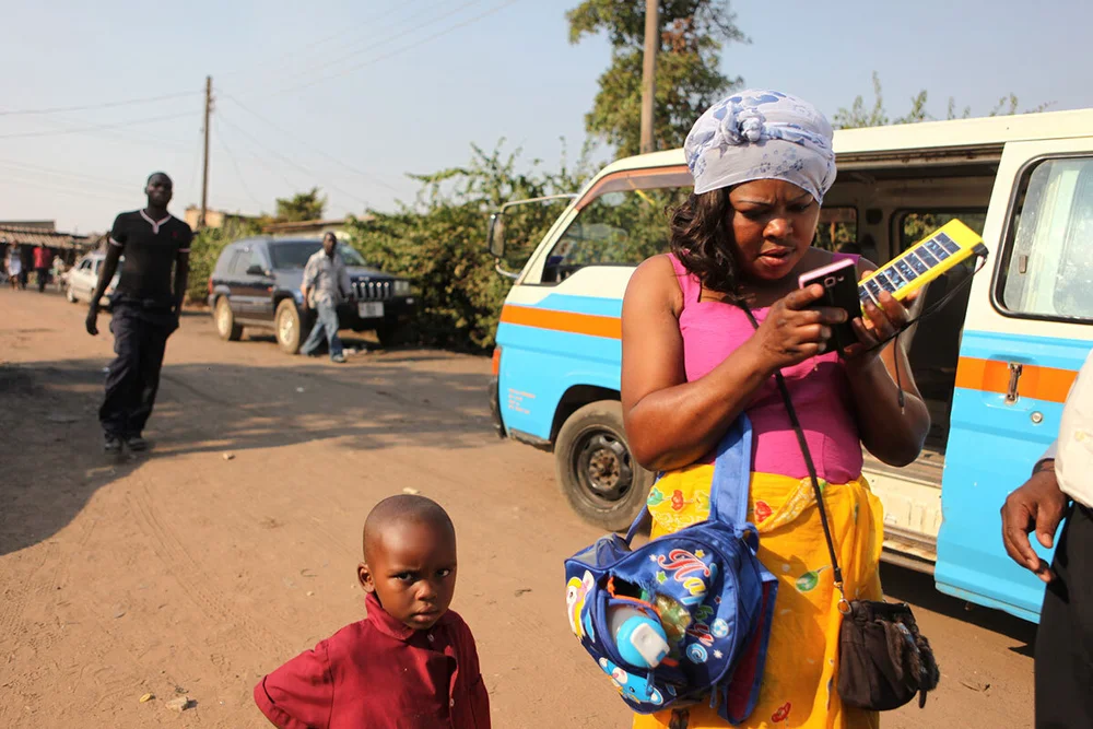A woman on a dirt road holding a phone cabled to a yellow handheld solar panel, a boy beside her and a van behind