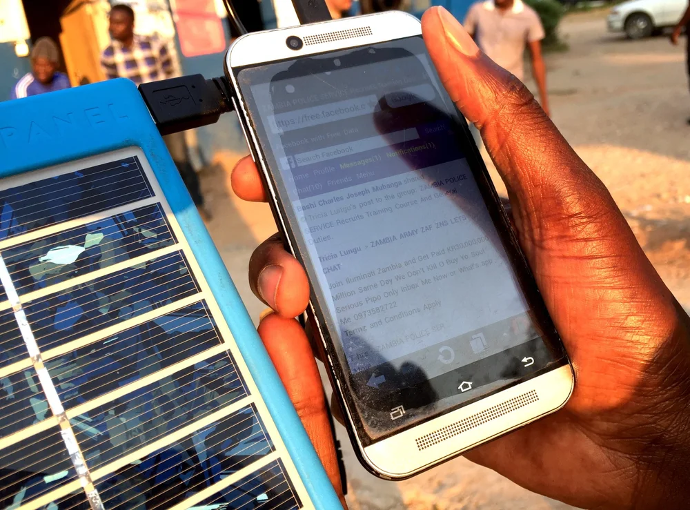 A hand holding a white phone showing Facebook with Free Data, connected by USB cable to a blue World Panel solar panel in sunlight