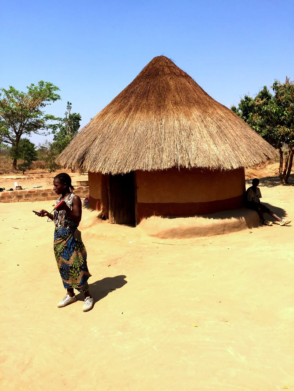 A young woman in a patterned wrap holding a phone and a small red device in a sandy yard before a thatched round hut