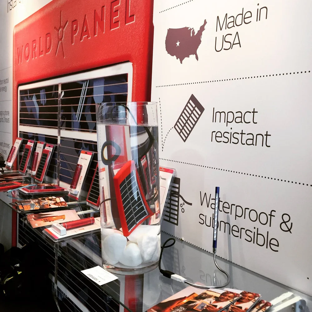 World Panel trade show display: a red solar panel submerged in a glass vase of water and white stones on a glass counter, beside packaged panels and a lit LED stick light