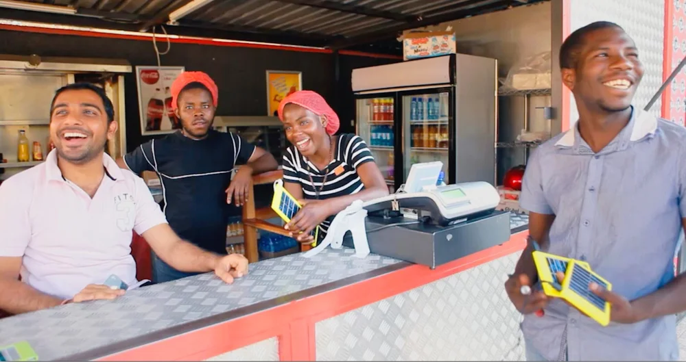 Four people laughing around a shop counter with yellow-cased World Panel solar devices, one held open like a folding panel, a cash register on the counter