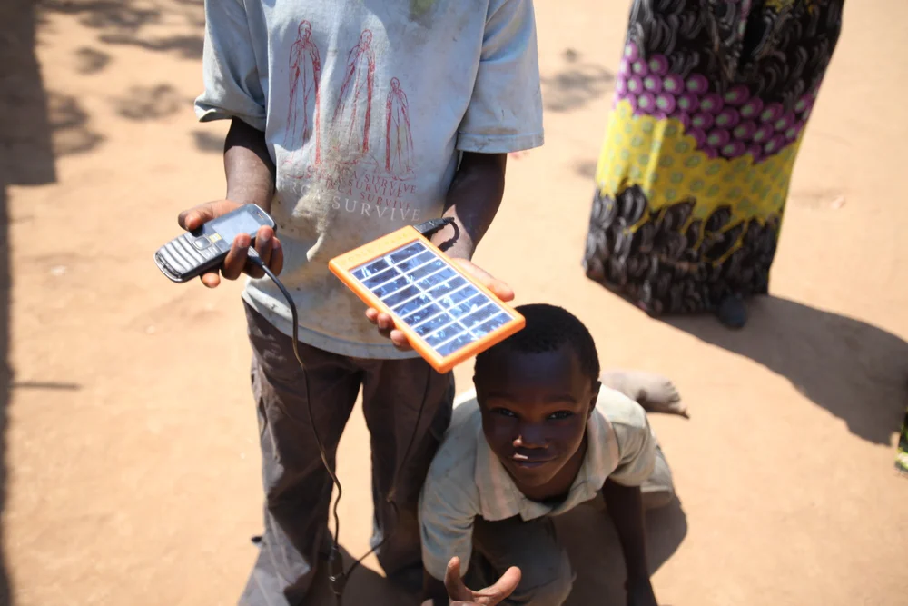 An overhead view of hands holding an orange World Panel solar panel cabled to a black phone, with a smiling boy sitting on the dirt below