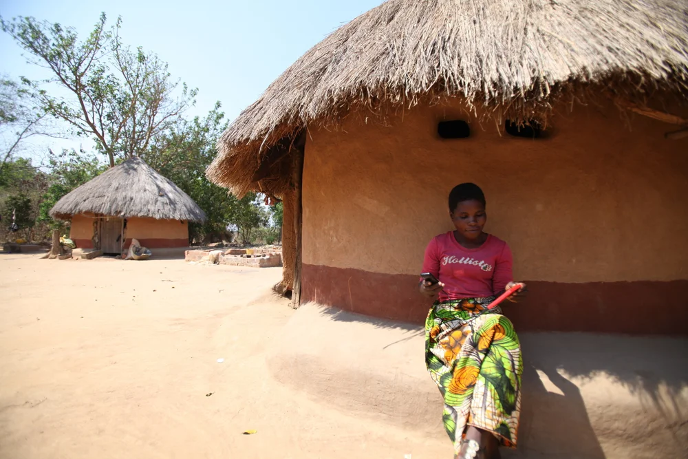 A young woman in a pink shirt sitting on the ledge of a thatched round hut, holding a phone in one hand and a red solar device in the other