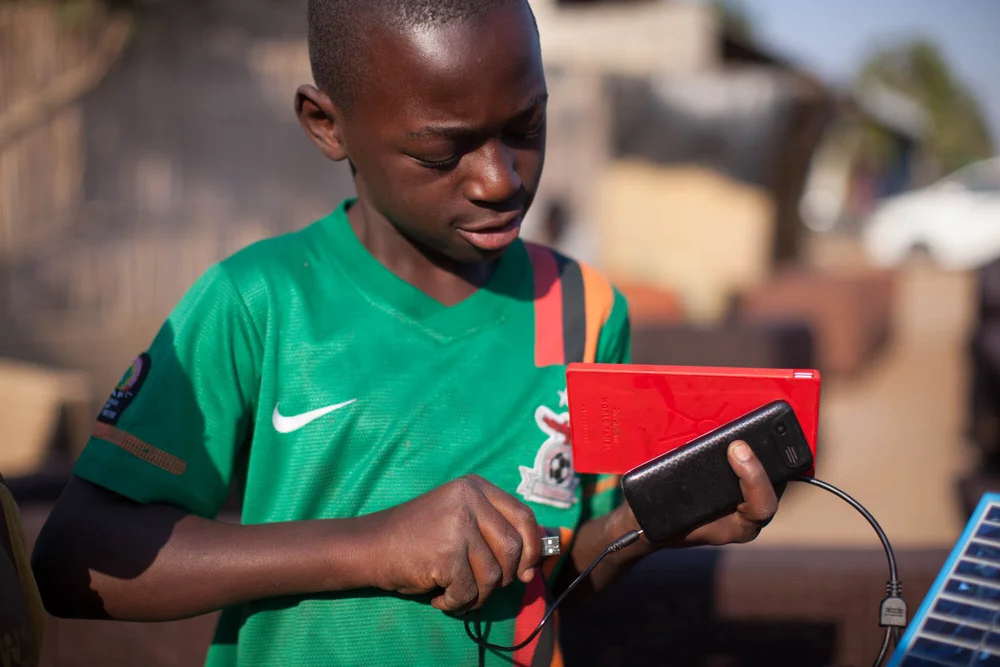 A boy in a green Zambia football jersey connecting a phone to a red World Panel SunStream device with a USB cable run from a mini solar panel