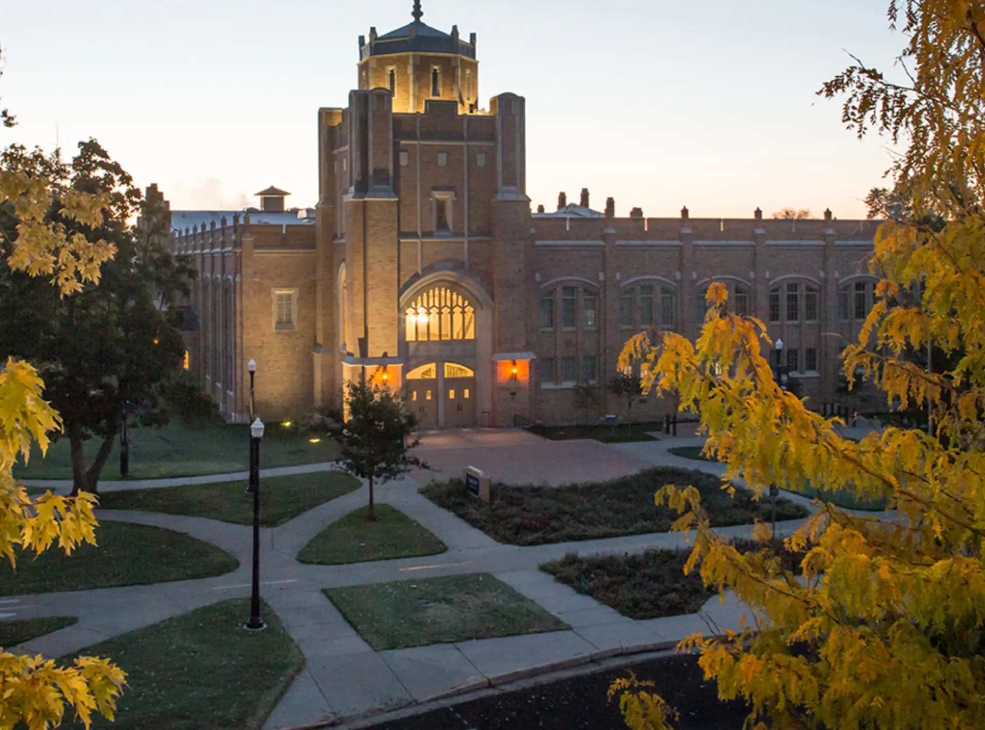 University of Northern Colorado campus at dusk: an illuminated stone tower and arched entrance framed by yellow autumn trees and crossing walkways