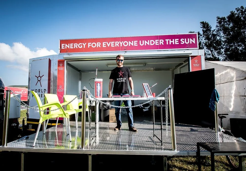 World Panel mobile trailer stage on a grassy field: “ENERGY FOR EVERYONE UNDER THE SUN” on a magenta banner above a table of solar panels and two lime-green chairs